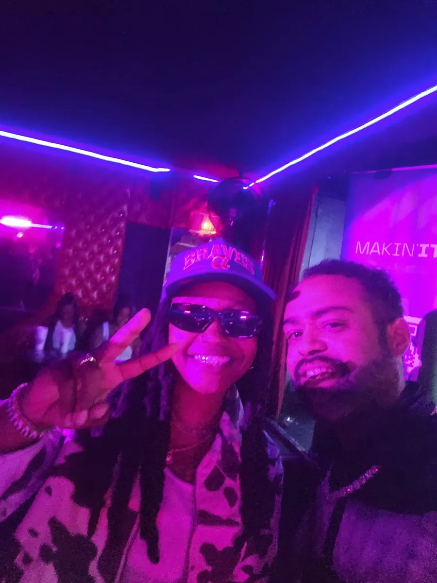 JC beside a guest in a cap and sunglasses flashing a peace sign, a MAKIN IT banner behind them at a BET weekend event