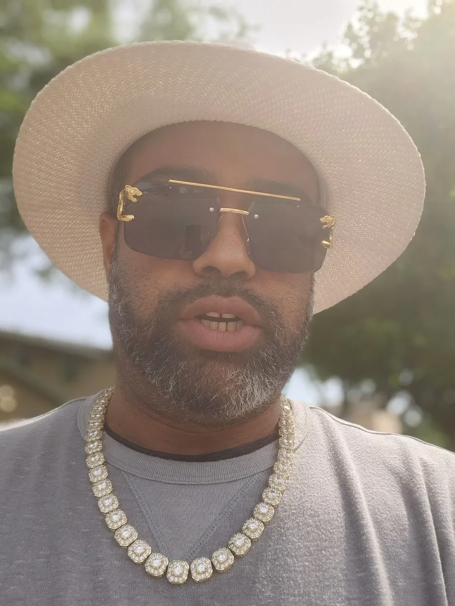 JC in a white wide-brim hat, gold-framed sunglasses, and a diamond chain, outdoors during BET Awards Weekend